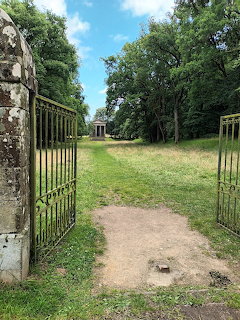 Open iron gates leading down a grass path to a small chapel surrounded by green trees