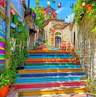 Rainbow-colored staircase climbing through a narrow sunny alley with plants and lanterns