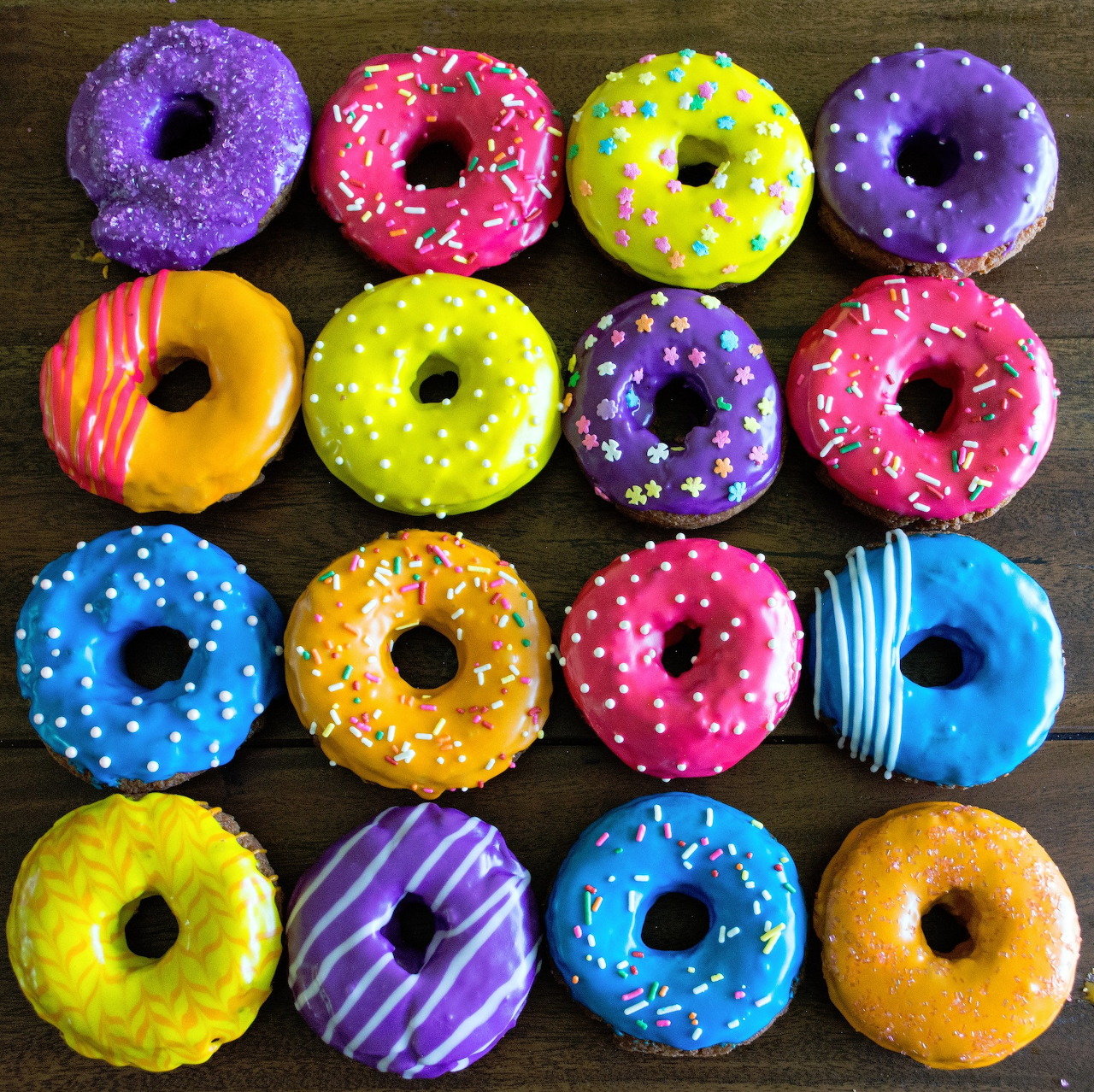 Sixteen colorful glazed donuts with various toppings arranged in a 4x4 grid on dark wood
