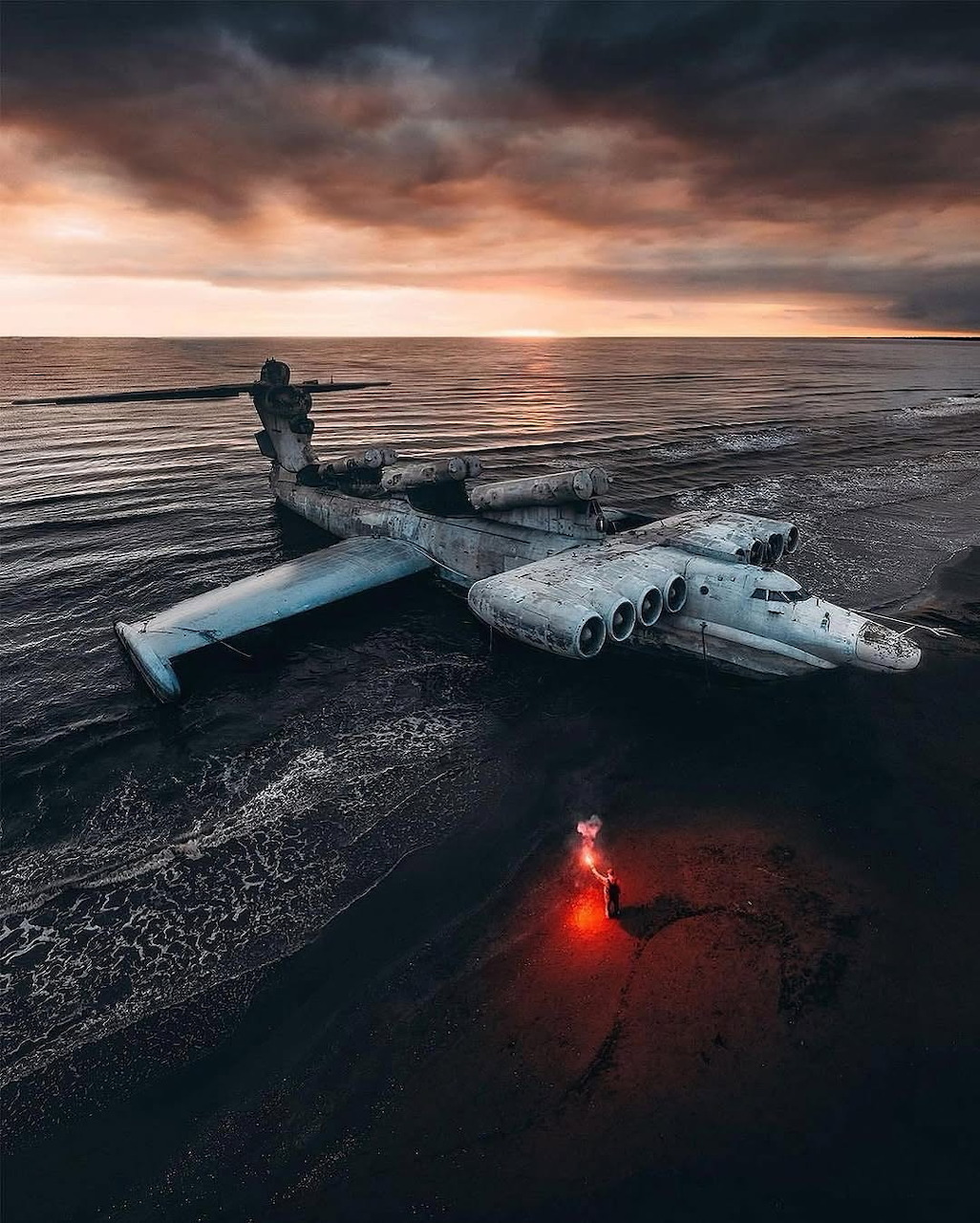 Aerial view of a giant abandoned Soviet ekranoplan on a beach at sunset with a person holding a red flare