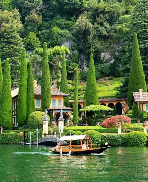 Lake Como villa with cone-shaped topiary trees, colorful buildings, and boat dock on green water with forested hillside