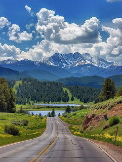 Road leading toward snow-capped mountains with green meadows, pine forests, and a lake under a cloudy blue sky