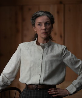 Woman in a white blouse standing indoors against a wooden wall
