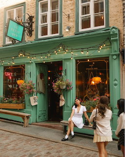 Two women sitting outside a green vintage café storefront decorated with string lights and flower baskets