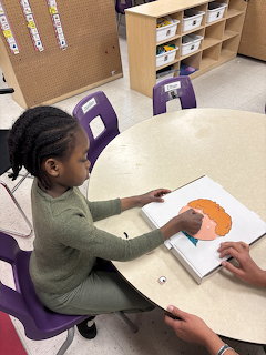 Young girl sitting at a round table in a classroom, coloring a drawing of a face