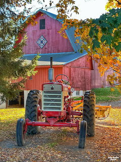 Vintage red tractor parked in front of a red barn with autumn trees and fallen leaves