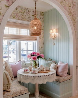 Pastel cozy nook with white table, tea set, pink roses, floral wallpaper and cushioned bench by window