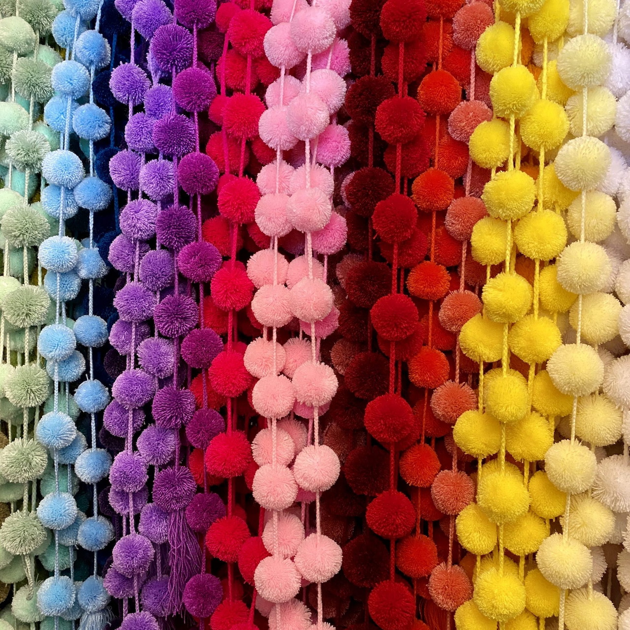 Vertical strands of fluffy yarn pom poms arranged in rainbow spectrum from blue through purple, pink, red, yellow to cream