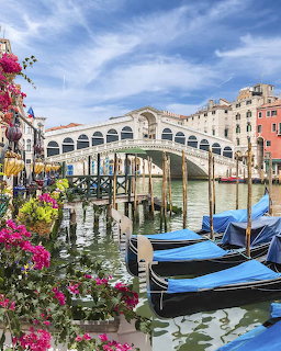 Boats on a Venice canal beneath a white arched bridge with flowers and historic buildings