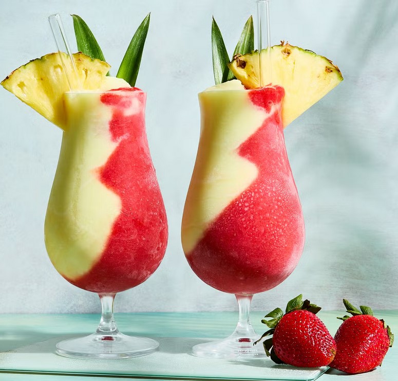 Two layered tropical cocktails with yellow and red swirls, pineapple garnish and fresh strawberries