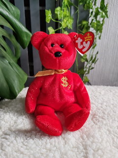 Red Ty Beanie Baby bear with gold ribbon and dollar sign, seated on a white textured surface