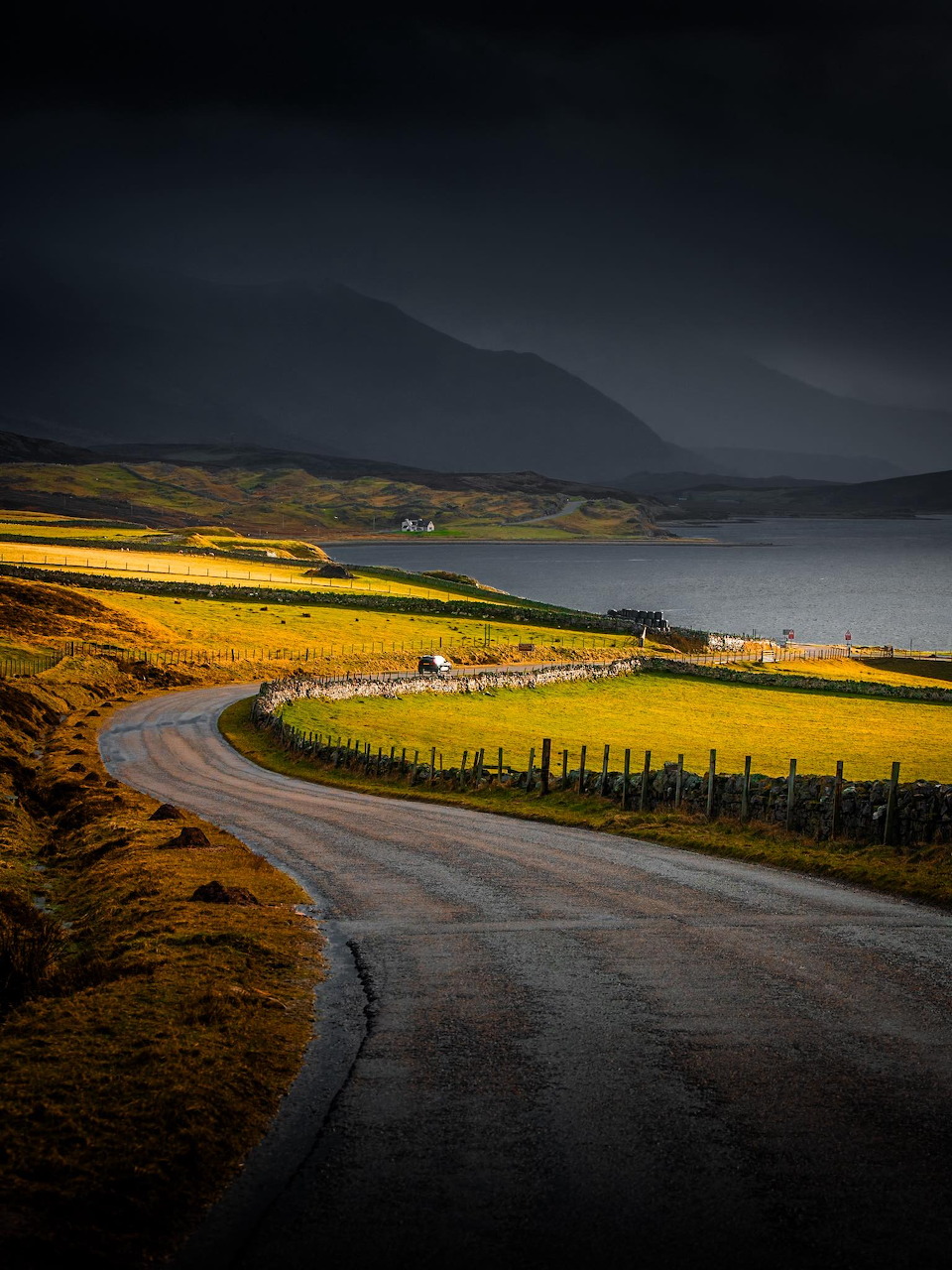 Winding road through golden fields along a Scottish loch with dark mountains and stormy sky
