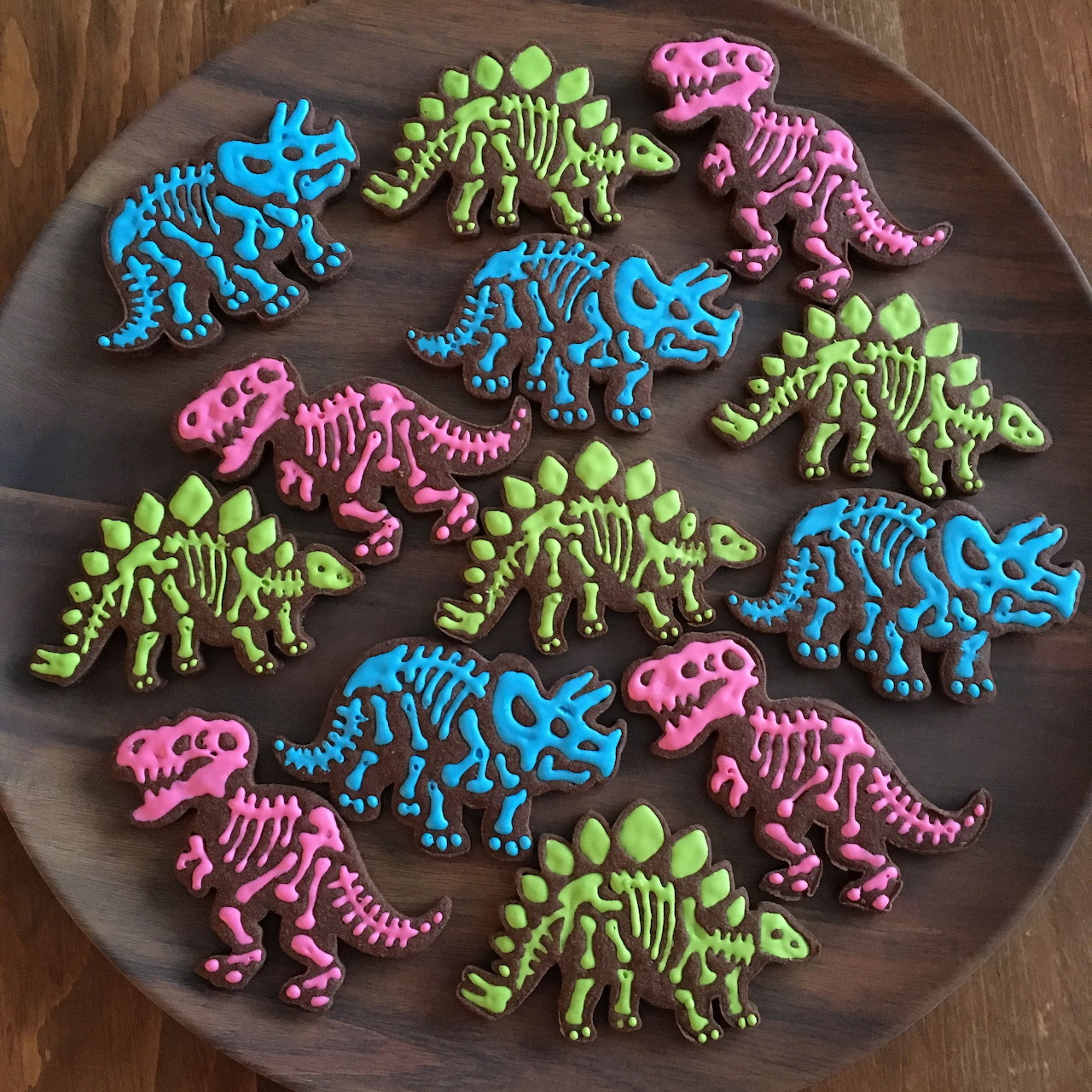 Assorted dinosaur skeleton cookies in bright pink, blue, and green icing arranged on a dark round plate