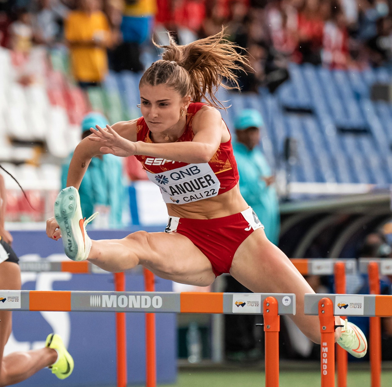 Female athlete jumping over hurdle in red uniform during track and field competition