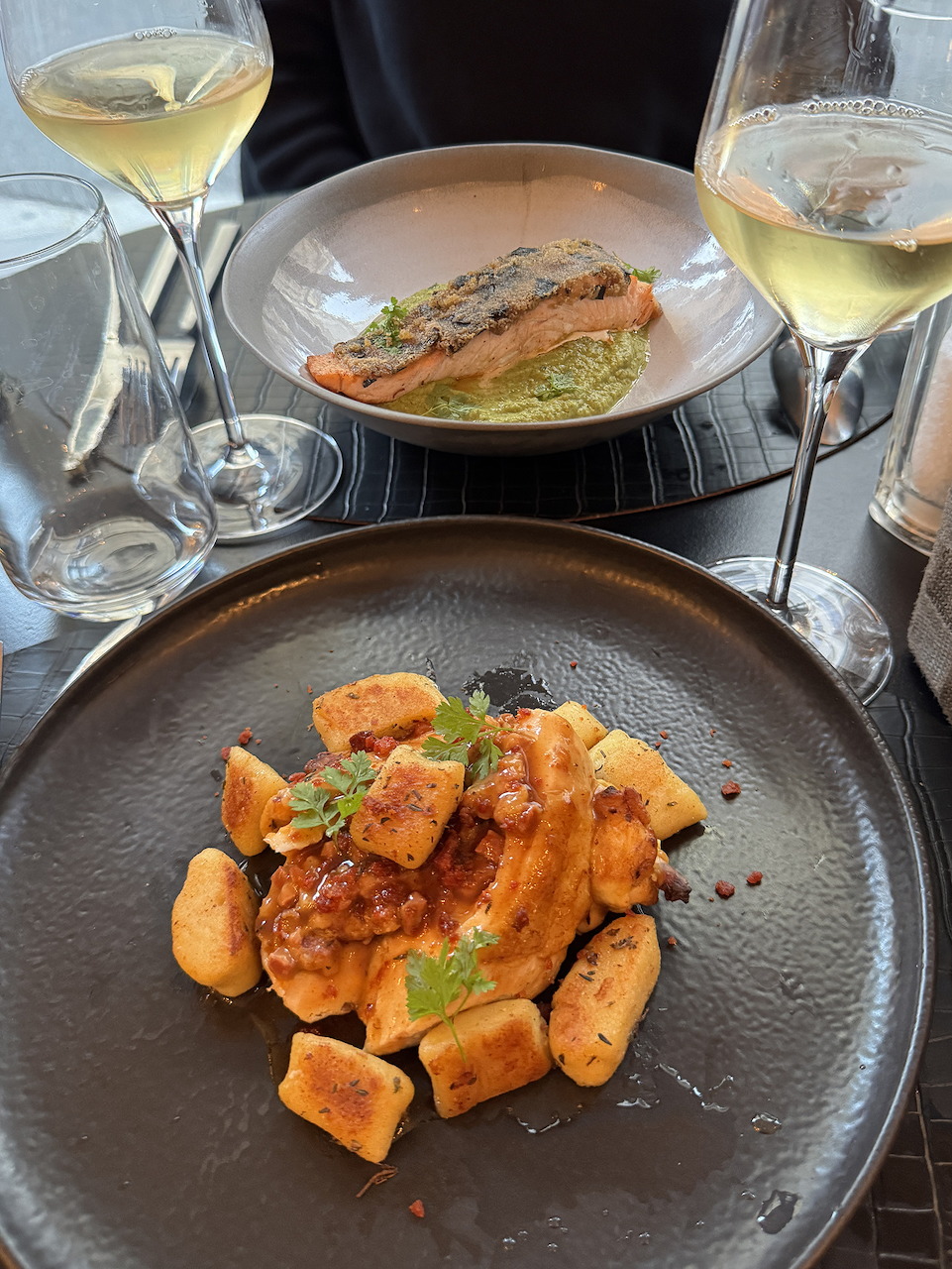 Restaurant table with gnocchi dish, fish fillet on green purée, and two glasses of white wine