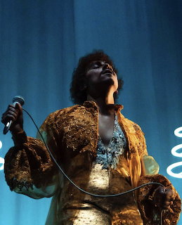 Singer on stage holding a microphone in a gold jacket under blue lighting
