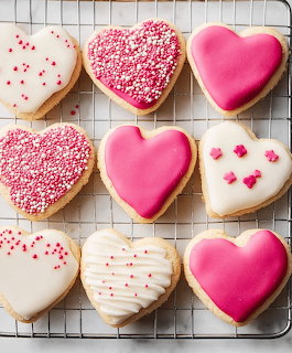 Nine heart-shaped cookies with pink and white frosting and red sprinkles on a wire cooling rack