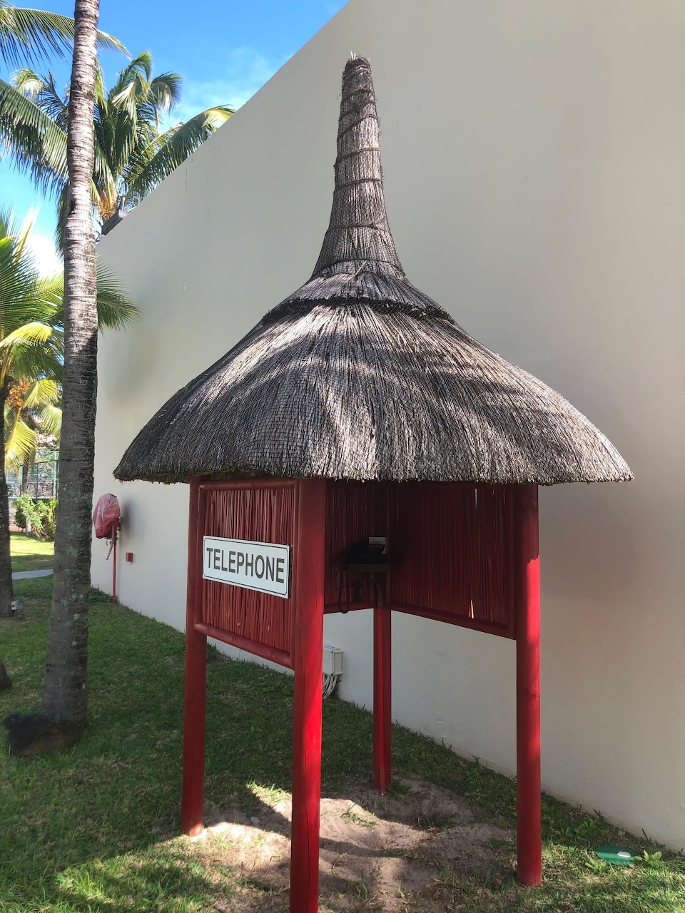 Small white phone booth with traditional thatched roof supported by red posts, palm trees beside it