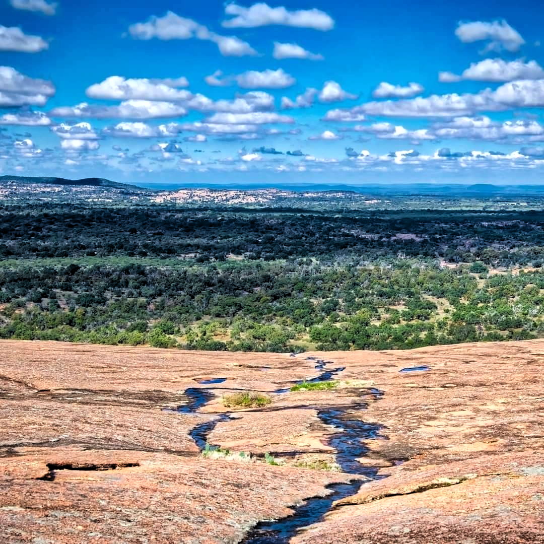 Wide desert landscape with sandstone foreground, green valley, and cloudy sky over distant mountains