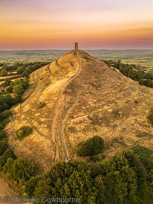 Aerial photo of Glastonbury Tor at sunset with St Michael's Tower atop a steep golden hill surrounded by trees