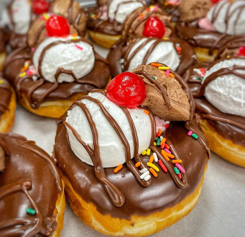 Rows of chocolate-covered donuts with whipped cream, chocolate drizzle, and maraschino cherry toppings