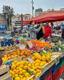 Outdoor fruit market stall with oranges, lemons, and bananas under red canopies with shopers