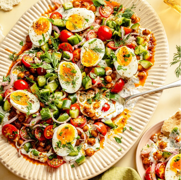 Oval platter of cucumber egg salad with boiled eggs, tomatoes, chickpeas, dill, and red onion on yellow tablecloth