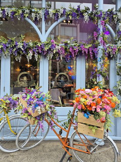 Two flower-decorated bicycles in front of a shop covered in purple wisteria and colorful blooms