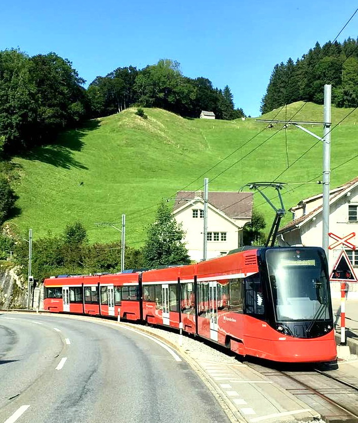 Modern red tram on a village road with green hills and blue sky in the background