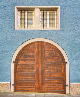 Arched wooden double door with louvers on a blue stucco building wall with a leaded glass window above
