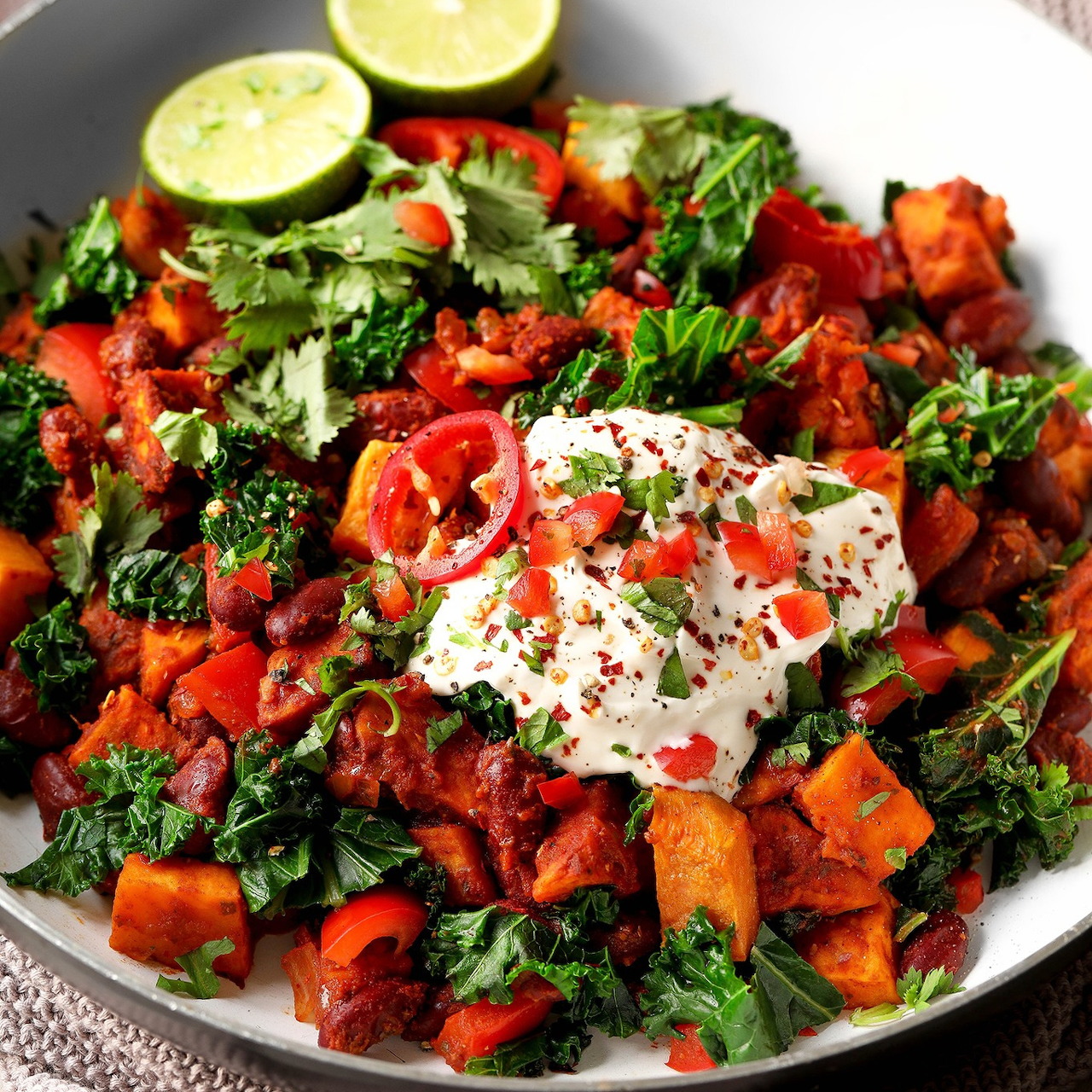 White bowl filled with roasted sweet potatoes, kale, red peppers, and yogurt, garnished with lime slices