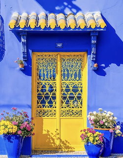 Ornate yellow door with intricate metalwork on a vivid blue wall flanked by flower pots