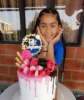 Smiling girl with braids posing behind a pink drip birthday cake with candy decorations and a golden topper