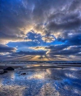 Dramatic clouds with sunbeams over a wet sandy beach at low tide with rocks and reflections