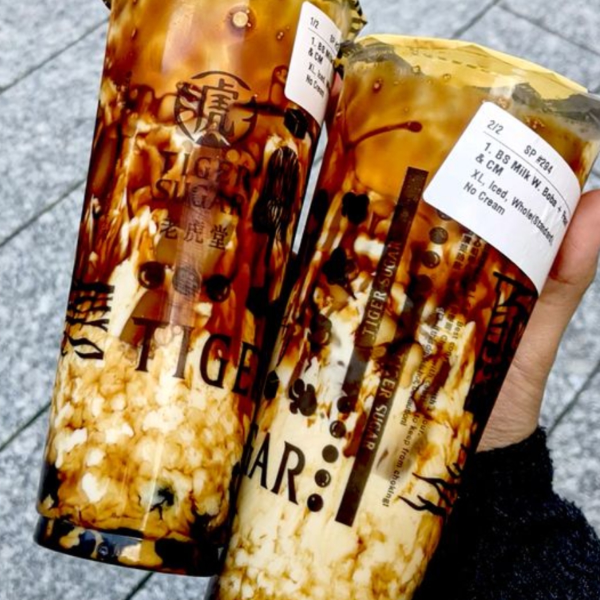 Two cups of brown sugar boba milk tea with tapioca pearls and cream being held by hand