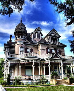 Grand Victorian mansion with turret and wraparound porch framed by trees against blue sky
