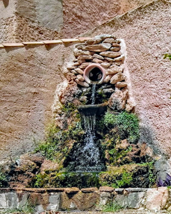 Rustic wall fountain with flowing water, stones, moss, and plants against a textured wall