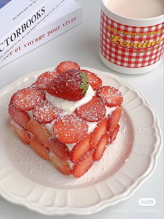 Heart-shaped strawberry cake with powdered sugar on white scalloped plate next to red gingham mug