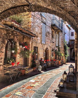 Sunny cobblestone street in Assisi with stone arch, cafe tables, flowers, and medieval stone buildings