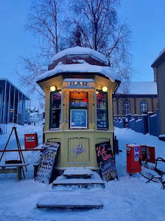 Yellow bar kiosk covered in snow with illuminated interior at dusk in a snowy city street