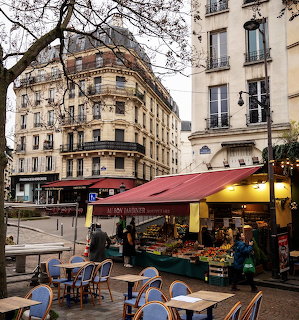 Paris street corner with a red awning market stall, cafe tables, and cream apartment buildings