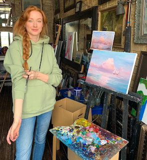 Woman with red braid in green hoodie standing in art studio next to paintings and palette