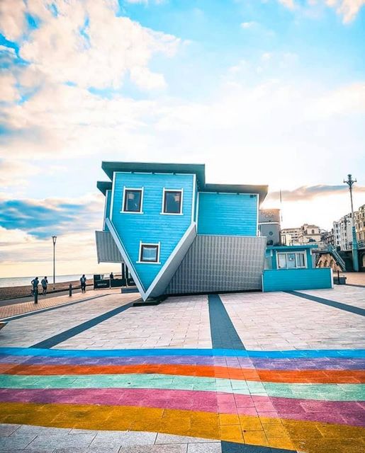 Blue upside-down house on a waterfront plaza with colorful striped pavement and bright sky