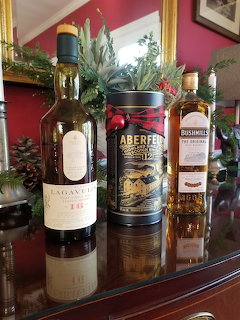 Three whiskey bottles on wooden table with holiday greenery and pine cones, red wall behind