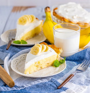Slice of banana cream pie with whipped cream, banana slices, and mint leaf beside a glass of milk