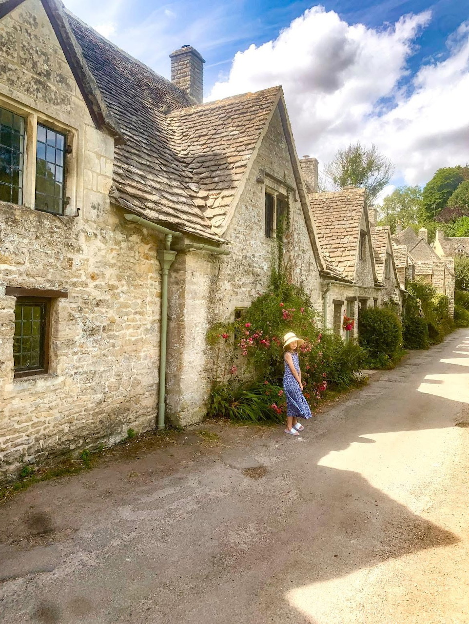 Person standing beside traditional Cotswolds stone cottages along a quiet village street under blue sky