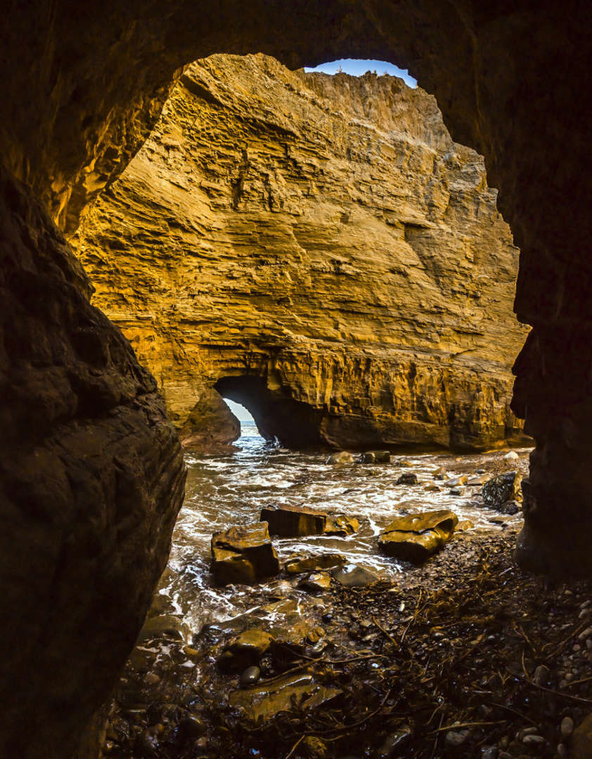 View through dark sea cave opening showing golden sunlit cliff face and rocky shore with tide pools