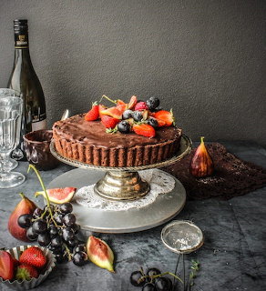 Dark chocolate tart topped with berries on a cake stand with figs, grapes, and a bottle nearby