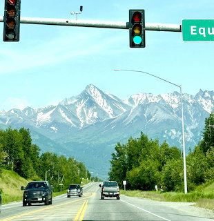 Road beneath traffic lights with green trees and snow-capped mountains in the background