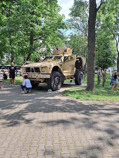 Tan military vehicle parked on a paved path in a tree-filled park with people nearby